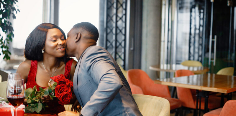Couple enjoying day out at restaurant. Black man gifted a bouquet of red roses for a woman. Woman wearing red elegant dress and man blue costume.