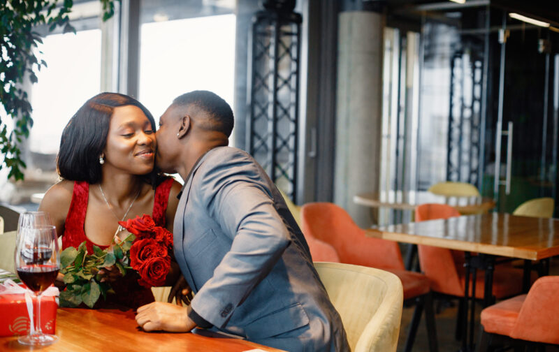 Romantic black couple sitting at restaurant wearing elegant clot Couple enjoying day out at restaurant. Black man gifted a bouquet of red roses for a woman. Woman wearing red elegant dress and man blue costume.